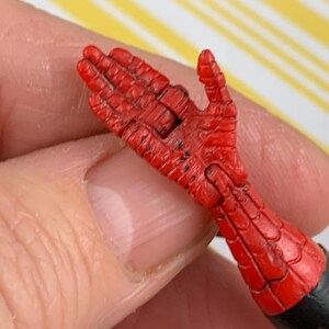 K&ouml;nnte beinhalten: Eine rote und schwarze Actionfigur-Hand mit einem strukturierten, netzartigen Muster. Die Hand ist mit leicht gekr&uuml;mmten Fingern dargestellt. Das Handgelenk ist schwarz, und die Hand wird von einer Person gehalten.