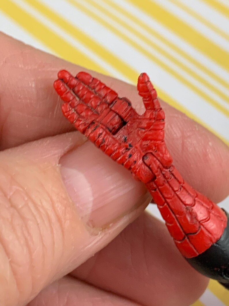 K&ouml;nnte beinhalten: Eine rote und schwarze Actionfigur-Hand mit einem strukturierten, netzartigen Muster. Die Hand ist mit leicht gekr&uuml;mmten Fingern dargestellt. Das Handgelenk ist schwarz, und die Hand wird von einer Person gehalten.