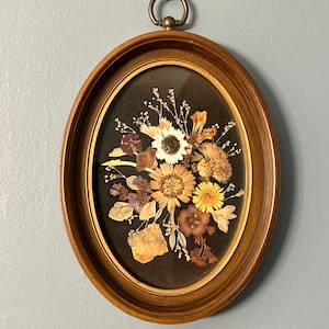 May include: Oval wooden framed pressed flower art. The frame is a light brown color with a brass ring for hanging. The pressed flowers are various colors including yellow, white, and brown, set against a black background.