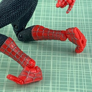 May include: A red and black action figure of Spider-Man, with articulated joints. The figure's legs and arms are red with a webbed design, while the torso and upper legs are black. The figure is posed on a green grid background.