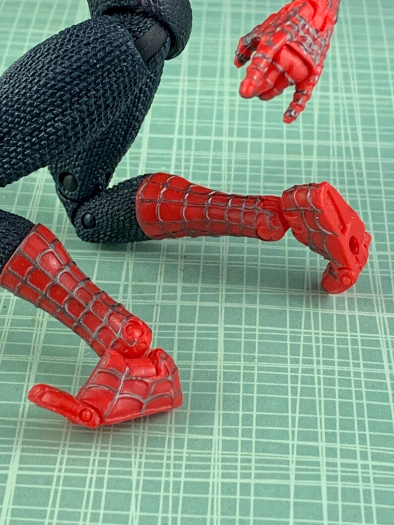 May include: A red and black action figure of Spider-Man, with articulated joints. The figure's legs and arms are red with a webbed design, while the torso and upper legs are black. The figure is posed on a green grid background.