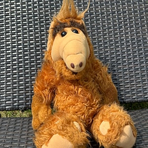 May include: A plush toy of the character Alf, featuring shaggy orange fur, a tan snout, and large black eyes. The toy is seated on a dark brown, woven surface, with its arms and legs extended.