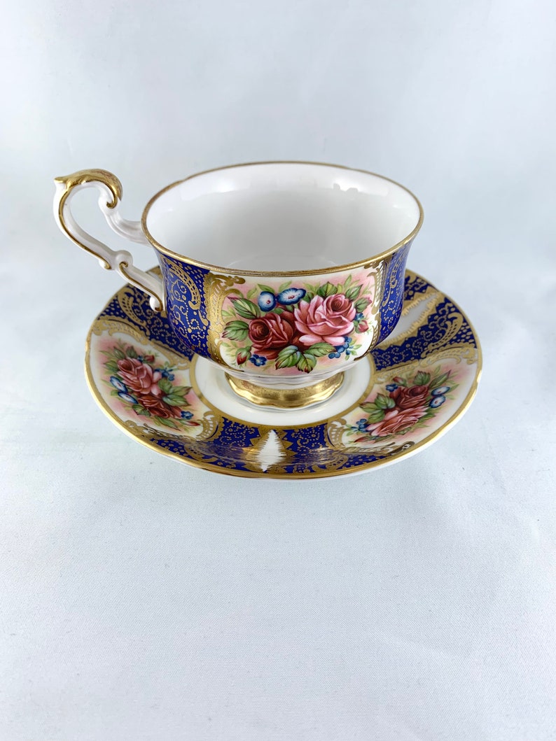 May include: A blue and gold teacup and saucer set with a floral design. The cup and saucer are decorated with a pink and red rose pattern.