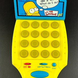 May include: A yellow handheld electronic game featuring a graphic of Homer Simpson with the text What do I do? What do I do? and The Simpsons Sez. The device includes a four-by-four grid of yellow buttons, a red power switch, a mode slider, and a blue start button.