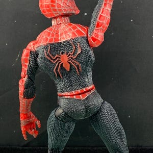 May include: A Spider-Man action figure in a dynamic pose, viewed from the back. The figure features a red and black color scheme with a spider emblem on the back. The figure's textured suit and web-like detailing are visible.