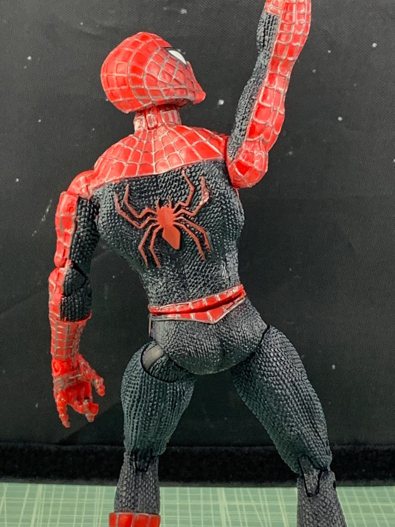 May include: A Spider-Man action figure in a dynamic pose, viewed from the back. The figure features a red and black color scheme with a spider emblem on the back. The figure's textured suit and web-like detailing are visible.