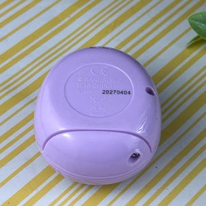 May include: A light purple, oval-shaped electronic device. The bottom of the device has the text "BANDAI WIZ B.13 CHINA" and the numbers "37480" and "29270404". The device has a small hole and a CE mark.
