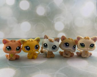 May include: A collection of five small cat figurines with oversized heads and large eyes. The cats are colored in shades of orange, yellow, white, and tan, each featuring distinct facial expressions and sitting in various poses on a flat surface.