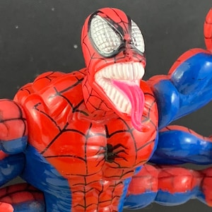 Peut inclure: Figurine articul&eacute;e de Spider-Man rouge et bleu avec une t&ecirc;te grotesque &agrave; bouche ouverte. La figurine a une longue langue rose, des dents blanches et des yeux en toile d'araign&eacute;e. Le corps est rouge avec des d&eacute;tails de toile noirs et des bras bleus.