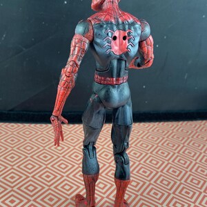 May include: A Spider-Man action figure, viewed from the back, showcasing a red and black costume. The figure's design includes web patterns on the arms and legs, with a spider emblem on the back. The figure is posed with arms extended.