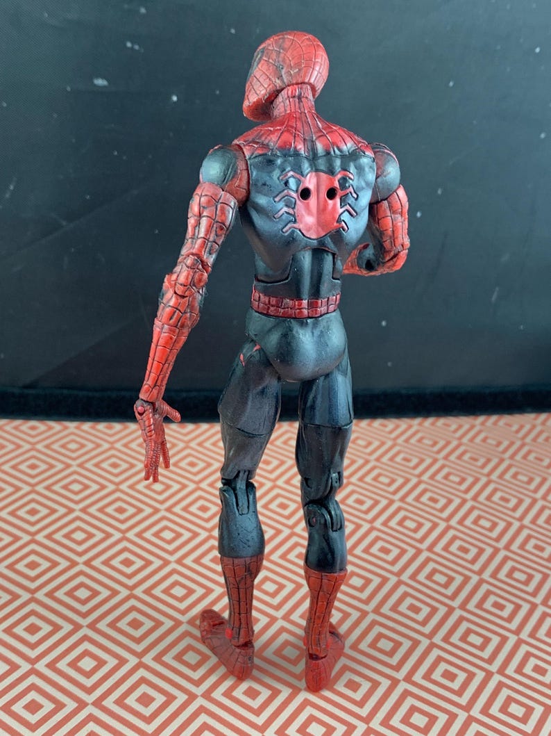 May include: A Spider-Man action figure, viewed from the back, showcasing a red and black costume. The figure's design includes web patterns on the arms and legs, with a spider emblem on the back. The figure is posed with arms extended.