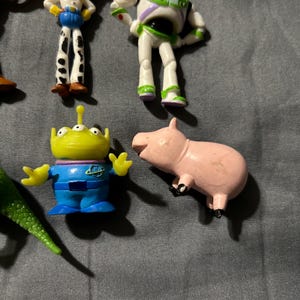Pu&ograve; includere: Una collezione di statuette dei personaggi di Toy Story, tra cui Buzz Lightyear, Jessie, un alieno e un maiale rosa. Le figure sono di plastica e dai colori vivaci. I giocattoli sono disposti su una superficie grigia.