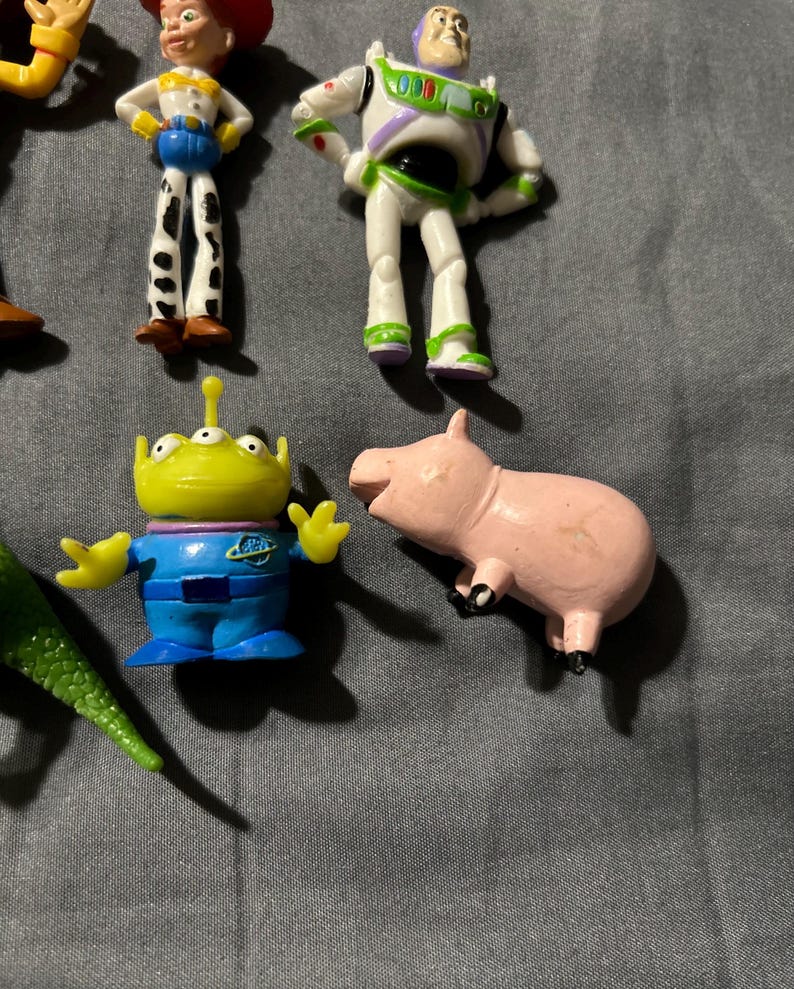 Pu&ograve; includere: Una collezione di statuette dei personaggi di Toy Story, tra cui Buzz Lightyear, Jessie, un alieno e un maiale rosa. Le figure sono di plastica e dai colori vivaci. I giocattoli sono disposti su una superficie grigia.