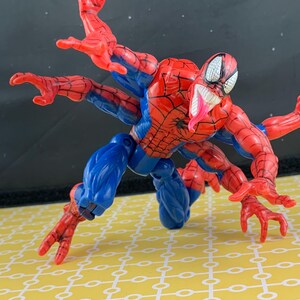 Peut inclure: Figurine articul&eacute;e de Spider-Man en position rampante, avec plusieurs bras. La figurine est principalement rouge et bleue, avec un motif de toile d'araign&eacute;e d&eacute;taill&eacute; sur le torse. Elle a une longue langue rose et des yeux blancs.