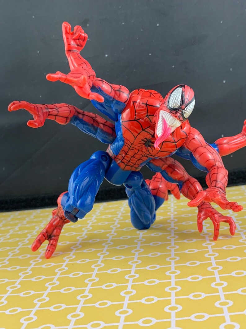 Peut inclure: Figurine articul&eacute;e de Spider-Man en position rampante, avec plusieurs bras. La figurine est principalement rouge et bleue, avec un motif de toile d'araign&eacute;e d&eacute;taill&eacute; sur le torse. Elle a une longue langue rose et des yeux blancs.