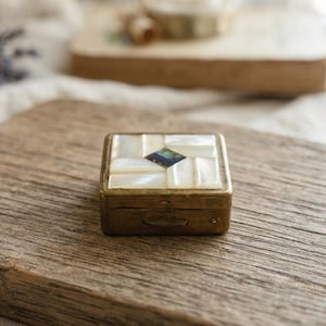 May include: A small square brass box featuring an inlaid lid with rectangular white mother-of-pearl tiles arranged in a geometric pattern around a single dark blue iridescent centre piece. The metal sides show a weathered, aged patina finish.