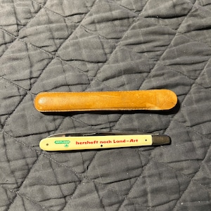 May include: A white folding pocket knife with a brown leather sheath. The knife has a red and green logo that says "Artland herzhaft nach Land-Art".