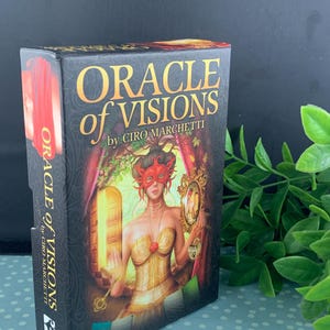 May include: A black box of "Oracle of Visions" cards by Ciro Marchetti. The box features a woman in a gold corset and red mask holding a key and a mirror. The text is in gold and white.
