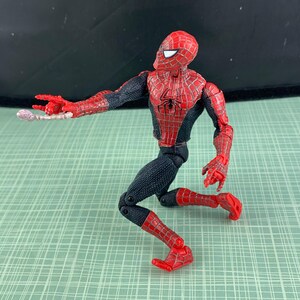 May include: A Spider-Man action figure in a crouching pose, featuring a red and black costume with web detailing. The figure has a red mask with white eye lenses and is holding a web-like accessory. The figure is on a green grid background.