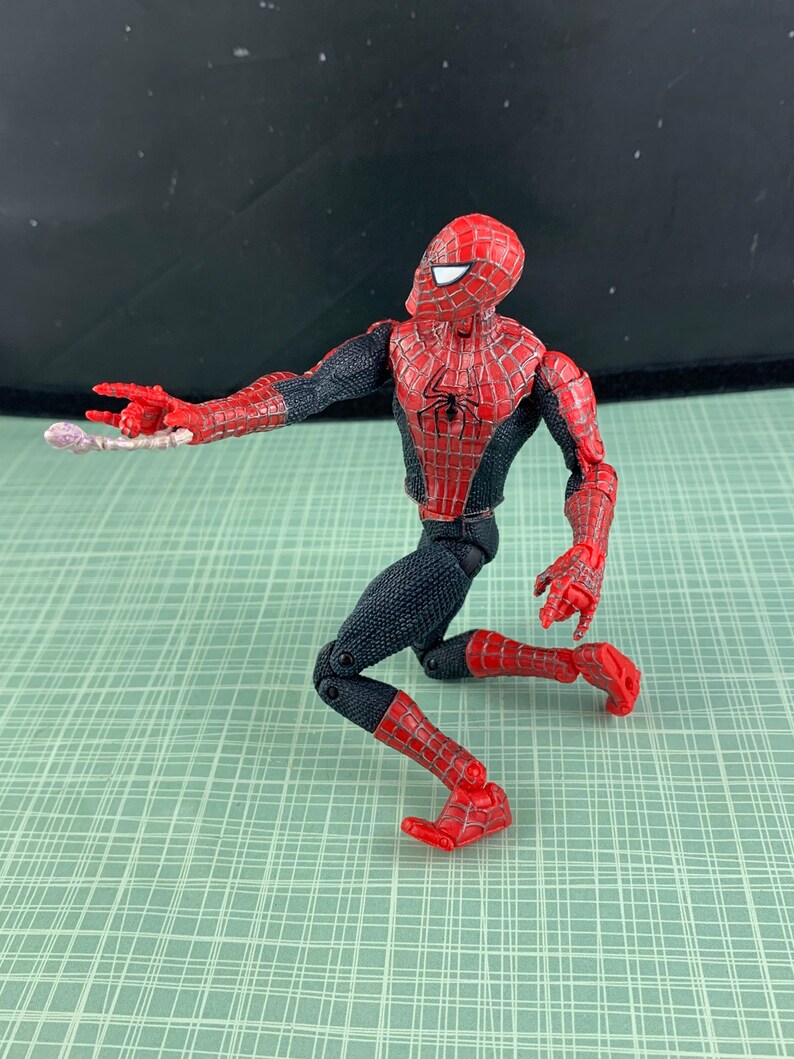 May include: A Spider-Man action figure in a crouching pose, featuring a red and black costume with web detailing. The figure has a red mask with white eye lenses and is holding a web-like accessory. The figure is on a green grid background.
