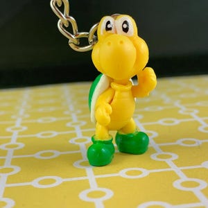 May include: A yellow Koopa Troopa keychain figure with a green shell and green shoes. The figure is attached to a silver chain. The background is a yellow surface with a white pattern.