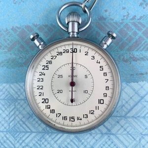 May include: A vintage stopwatch with a silver metal casing and a white face. The face displays black numerals and markings, with a red second hand. The stopwatch is attached to a metal chain and ring.