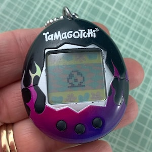 Op de afbeelding: Een zwarte en paarse Tamagotchi met een zilveren rand en een scherm dat een gepixelde afbeelding weergeeft. De Tamagotchi heeft drie knoppen aan de onderkant en de tekst "Tamagotchi" staat bovenaan gedrukt.