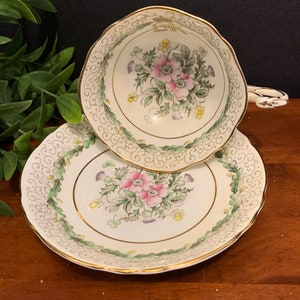 K&ouml;nnte beinhalten: Ein Vintage-Teetassen- und Untertassen-Set mit einem Blumendesign. Tasse und Untertasse sind wei&szlig; mit gr&uuml;nem und goldenem Rand. Das Blumendesign zeigt rosa und gelbe Blumen mit gr&uuml;nen Bl&auml;ttern.