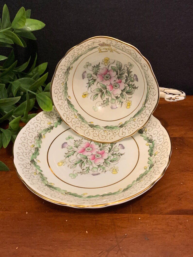 K&ouml;nnte beinhalten: Ein Vintage-Teetassen- und Untertassen-Set mit einem Blumendesign. Tasse und Untertasse sind wei&szlig; mit gr&uuml;nem und goldenem Rand. Das Blumendesign zeigt rosa und gelbe Blumen mit gr&uuml;nen Bl&auml;ttern.