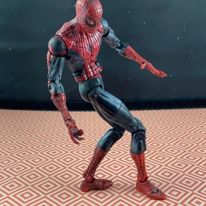 May include: A Spider-Man action figure in a dynamic pose. The figure features a red and black color scheme, with a detailed web pattern on the red sections. The figure is articulated and stands on a patterned surface.