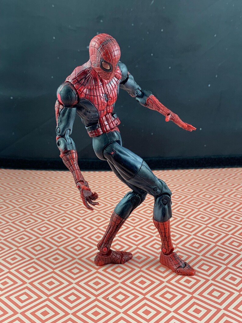 May include: A Spider-Man action figure in a dynamic pose. The figure features a red and black color scheme, with a detailed web pattern on the red sections. The figure is articulated and stands on a patterned surface.