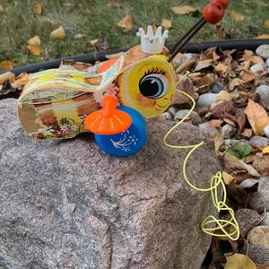 May include: A vintage wooden pull toy shaped like a bee with a yellow and brown body, orange wings, and a white crown. The bee has large blue eyes and a smiling face. The toy has a yellow pull string.