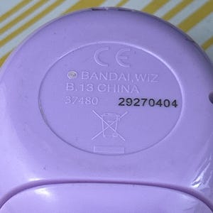 May include: Close-up of a light purple plastic device with the text "CE", "BANDAI, WIZ", "B.13 CHINA", and the numbers "37480" and "29270404". A crossed-out trash can symbol is also visible.