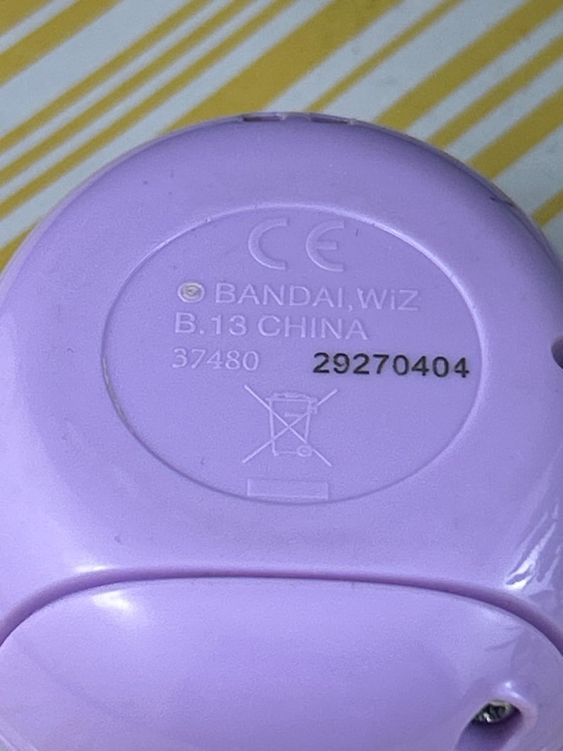 May include: Close-up of a light purple plastic device with the text "CE", "BANDAI, WIZ", "B.13 CHINA", and the numbers "37480" and "29270404". A crossed-out trash can symbol is also visible.