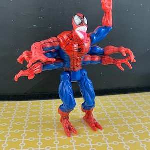 Peut inclure: Figurine articul&eacute;e de Spider-Man, en rouge et bleu, dans une pose dynamique. Elle poss&egrave;de plusieurs bras, un motif de toile d'araign&eacute;e d&eacute;taill&eacute; et une langue tir&eacute;e. La figurine est pos&eacute;e sur une surface jaune &agrave; motifs.