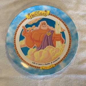 May include: Round plate featuring Disney's Hercules. The plate has a blue rim with a decorative border. The center depicts Zeus in orange and purple, holding a lightning bolt. Text reads "Life is just a bowl of chariots!" and "Zeus."