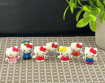 May include: A selection of small Hello Kitty figurines. Each figurine is white with a different coloured bow and outfit. The outfits include pink, blue, red, orange, and yellow. One figurine is dressed as a sailor.