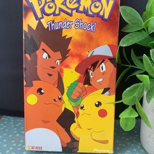 May include: A VHS tape for "Pokémon: Thunder Shock!" features the Pokémon logo in yellow and blue. The cover art shows characters from the animated series, including Pikachu and Ash Ketchum, against a fiery background. The tape is from VIZ Video.