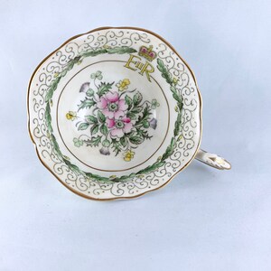 K&ouml;nnte beinhalten: Eine wei&szlig;e Teetasse mit goldfarbenem Rand und einem floralen Design. Das Design zeigt eine rosa Blume, gr&uuml;ne Bl&auml;tter und eine gelbe Krone mit den Buchstaben "E.R." in Gold.