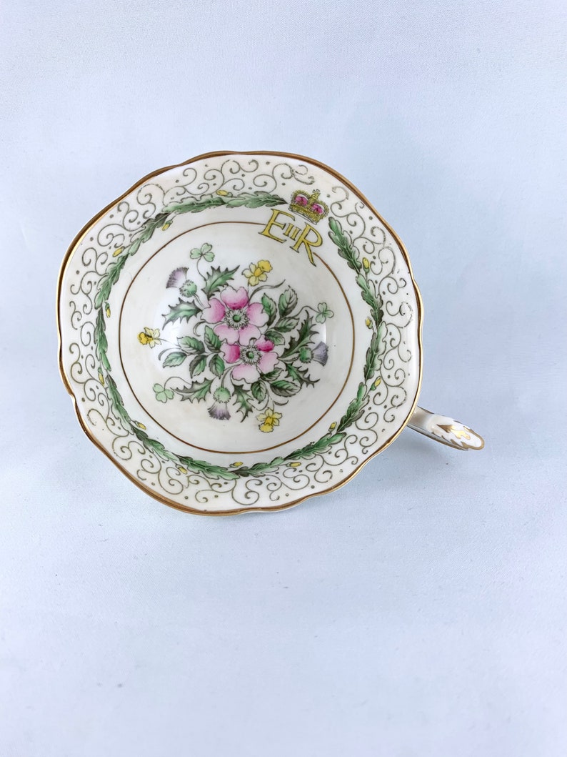 K&ouml;nnte beinhalten: Eine wei&szlig;e Teetasse mit goldfarbenem Rand und einem floralen Design. Das Design zeigt eine rosa Blume, gr&uuml;ne Bl&auml;tter und eine gelbe Krone mit den Buchstaben "E.R." in Gold.