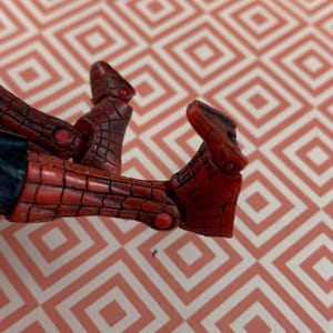 May include: Close-up of a red and black action figure's feet. The figure's feet are detailed with a textured pattern and small red accents. The figure is positioned against a patterned background.