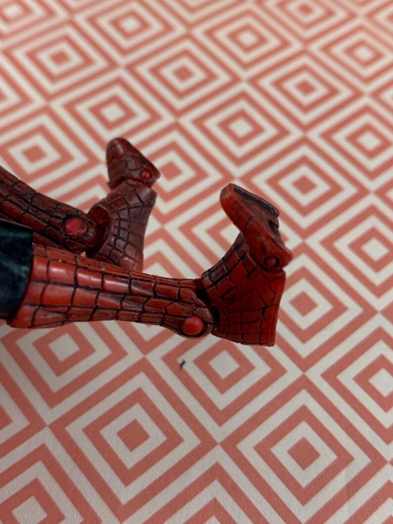 May include: Close-up of a red and black action figure's feet. The figure's feet are detailed with a textured pattern and small red accents. The figure is positioned against a patterned background.