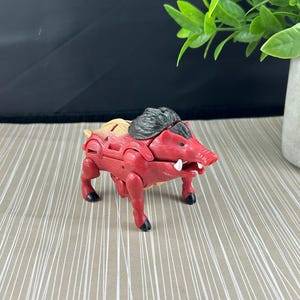 以下が含まれることがあります： 黒い蹄と黒いタテガミを持つ、赤とタン色の玩具イノシシ。おもちゃは白い牙を持ち、模様のある表面に置かれています。おもちゃはプラスチック製の置物です。