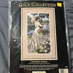 以下が含まれることがあります： DimensionsのThe Gold Collectionによる「A Summer Stroll」というタイトルのクロスステッチキット。パッケージには、日傘とバスケットを持って庭にいる長いドレスを着た人物のイラストが描かれています。フレームサイズは25x46cmと記載されています。