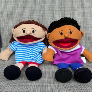 Peut inclure: Deux marionnettes à main en peluche avec des cheveux en laine marron et noir. La marionnette de gauche porte un t-shirt rayé bleu et blanc et un short rouge. Celle de droite porte un t-shirt violet à manches blanches et un pantalon bleu.
