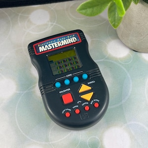 May include: A black electronic handheld Mastermind game. The game has a screen displaying colored pegs, with buttons for code entry, scrolling, and game functions. The text "MASTERMIND" is displayed at the top.