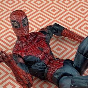 May include: A Spider-Man action figure in a dynamic pose, featuring a red and black costume with web detailing. The figure is lying on a patterned surface with a geometric design. The figure's eyes are white.