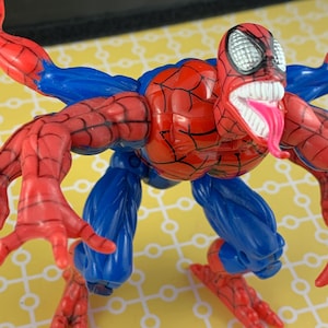 Peut inclure: Figurine articul&eacute;e de Spider-Man, rouge et bleu, avec plusieurs bras et jambes. Le personnage a une bouche ouverte avec des dents et une longue langue rose, avec des d&eacute;tails de toile d'araign&eacute;e.