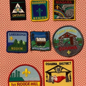 May include: A collection of embroidered patches featuring various designs and text. Patches include the words "Ontario," "Niagara Region," "Lakeridge Region," "North Waterloo," "Camp Samac Oshawa," "1st Rouge Hill," and "Oshawa District 80th Anniversary."