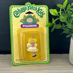 Peut inclure: Une figurine Cabbage Patch Kids dans son emballage d'origine. La poupée aux cheveux jaunes porte une robe blanche avec des bordures roses et un chapeau de fête. La carte présente des bordures vertes, des rayures jaunes et le texte Cabbage Patch Kids FIGURINE, MADE IN CHINA.